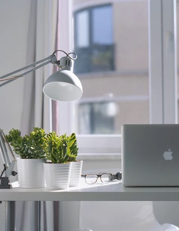 a photo of a table near a window with a laptop & a plant and a lamp beside it